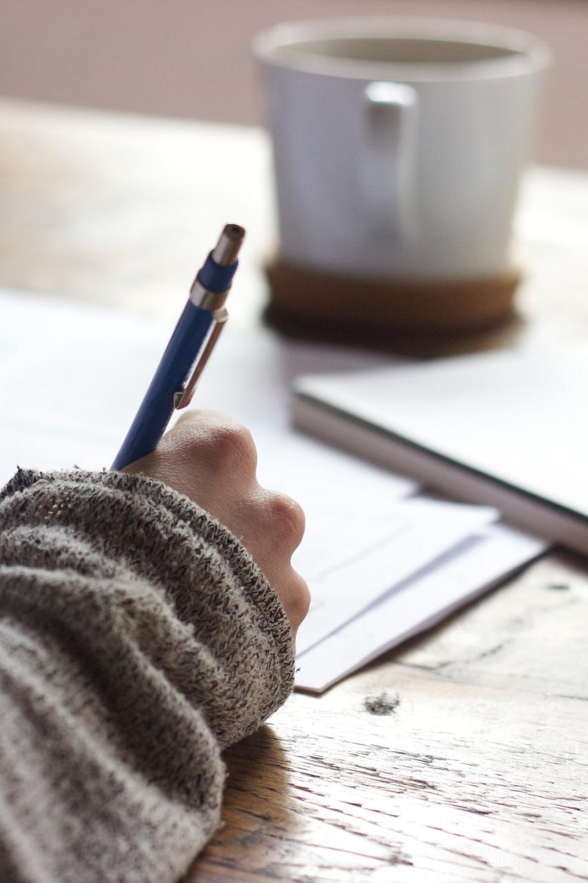 person writing on brown wooden table near white ceramic mug